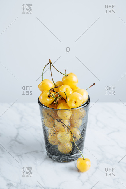 From above of full glass of fresh yellow cherries with stalks on marble table against white background
