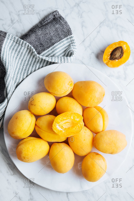 Top view of white plate with fresh yellow ripe apricots placed on plate near table cloth on white marble table with cut in half apricot