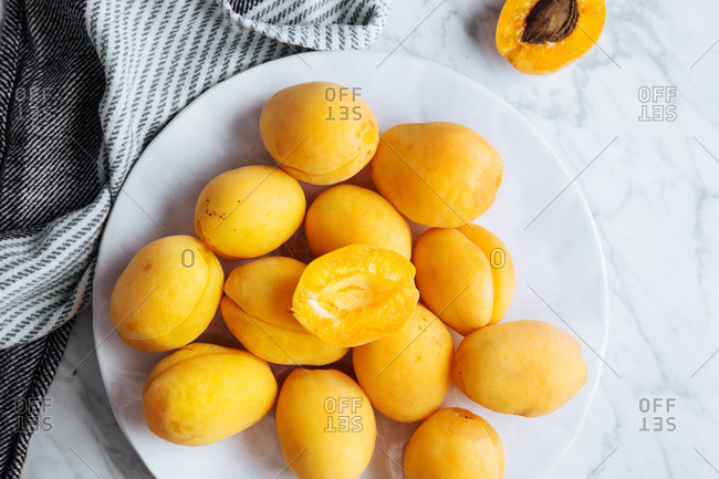Top view of white plate with fresh yellow ripe apricots placed on plate near table cloth on white marble table with cut in half apricot