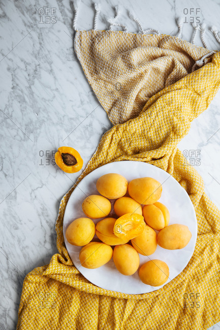 Top view of white plate with fresh yellow ripe apricots placed on yellow cloth on white marble table with cut in half apricot