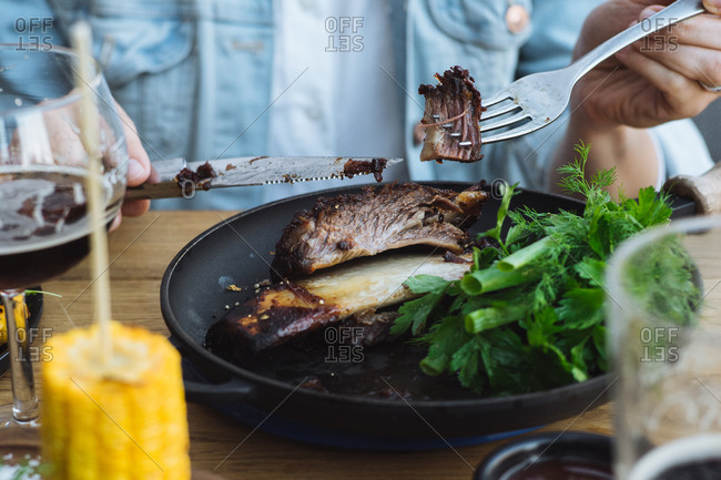 Crop unrecognizable person with knife and fork eating delicious grilled beef steak with bone served with fresh greenery and grilled corn in restaurant