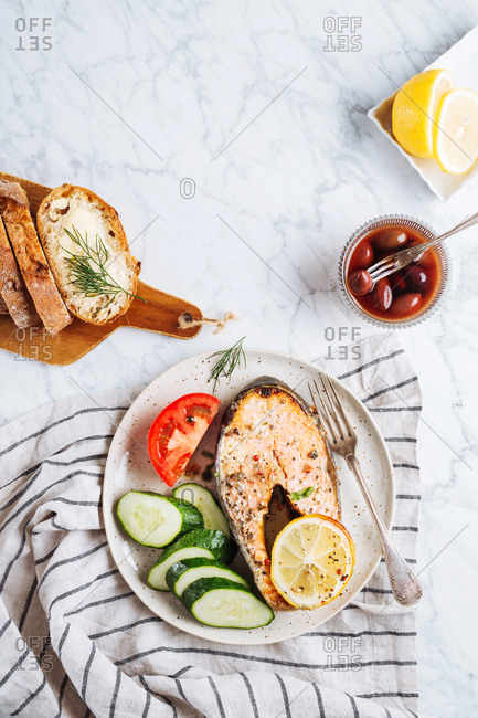 Top view of delicious healthy dinner with aromatic grilled salmon steak with lemon and herbs served with fresh sliced cucumber and tomato on marble table with fresh bread and olives