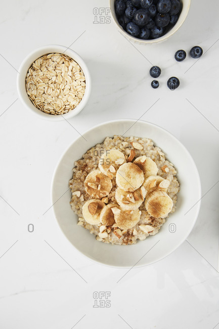 Bowl of porridge topped with banana,  almonds and cinnamon,  a bowl of oats and a  bowl of blueberries on white marble countertop