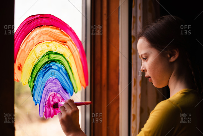 Young girl painting rainbows on a window to support Stay-at-Home