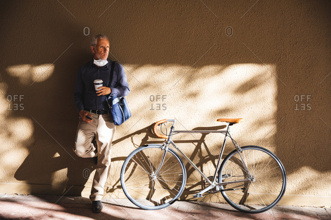 Senior Caucasian man out and about in the city streets during the day, wearing a face mask against coronavirus, covid 19, leaning on the wall and holding a cup of takeaway coffee while his bicycle is standing next to him.