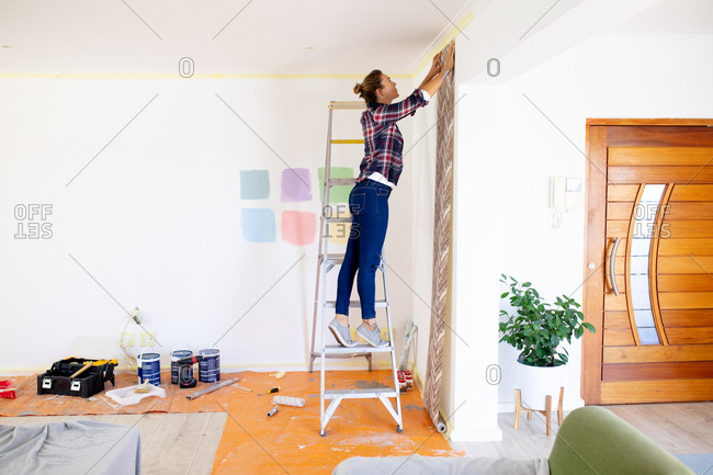 Caucasian woman spending time at home self isolating and social distancing in quarantine lockdown during coronavirus covid 19 epidemic, posing wallpaper standing on a ladder.