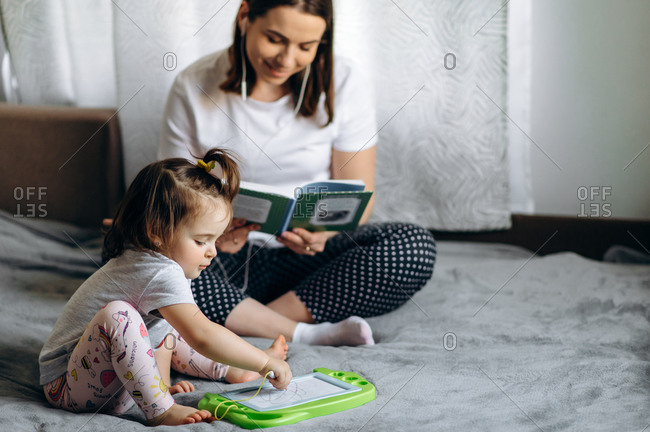 Adorable little baby girl draws on a magnetic board sitting on a sofa at home, and her mother sits in the background reading a book and listening to music