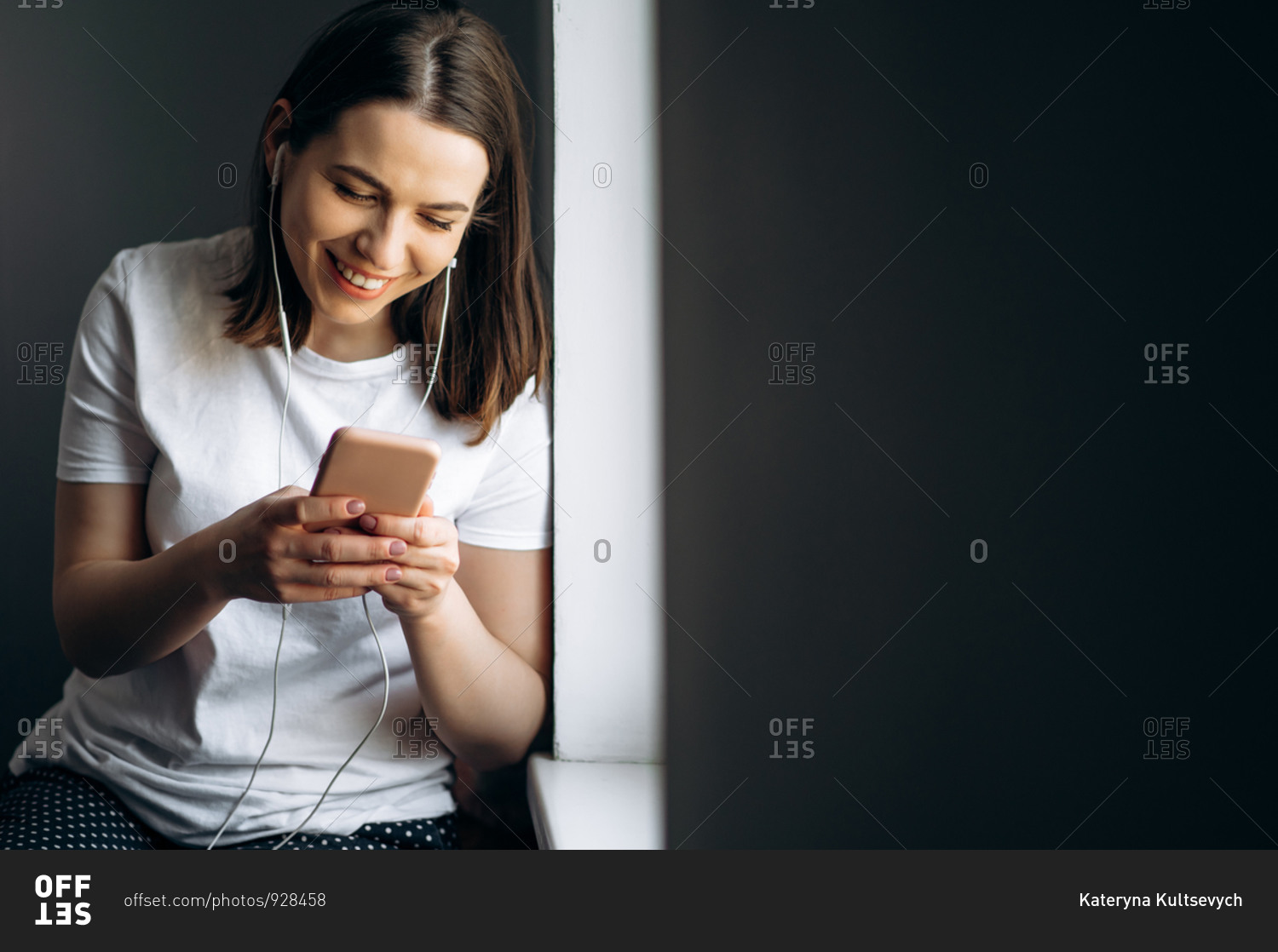 Attractive young girl listens to her favorite music on the phone via headphones near the window at home, chatting online and smiling