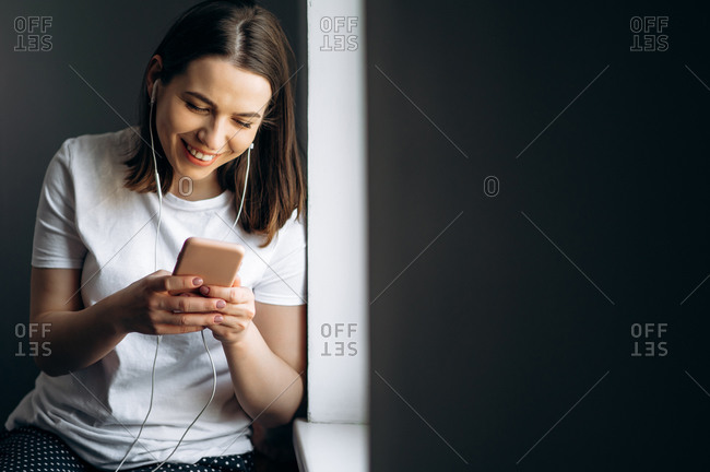 Attractive young girl listens to her favorite music on the phone via headphones near the window at home, chatting online and smiling