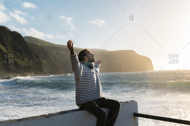 Man sitting at the coast at sunset with outstretched arms- Sao Miguel Island- Azores- Portugal