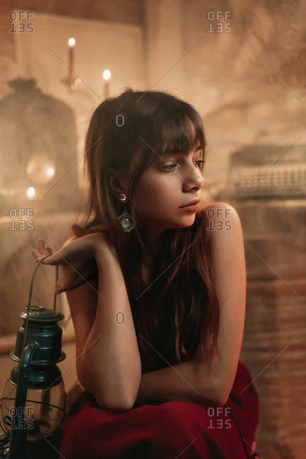 Portrait of a young woman in her twenties wearing a long red dress sitting and holding an old blue hand lamp inside her studio with candles and fog in the air.
