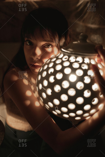 Portrait of a young woman in her twenties holding a perforated round ceramic lamp with a bulb light.
