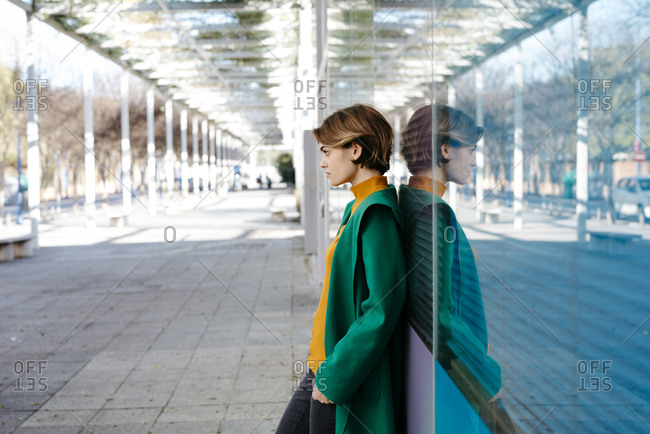 Portrait of young woman wearing green coat and leaning on colorful glass wall with her reflection