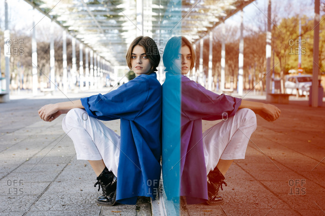 Young woman with urban look sitting on the floor and leaning on colorful glass wall with her reflection