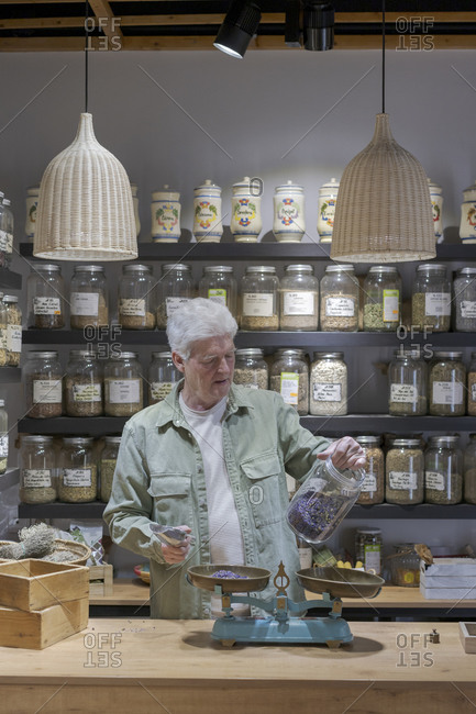 Senior man behind the counter in his shop holding jar of dried lavender blossoms