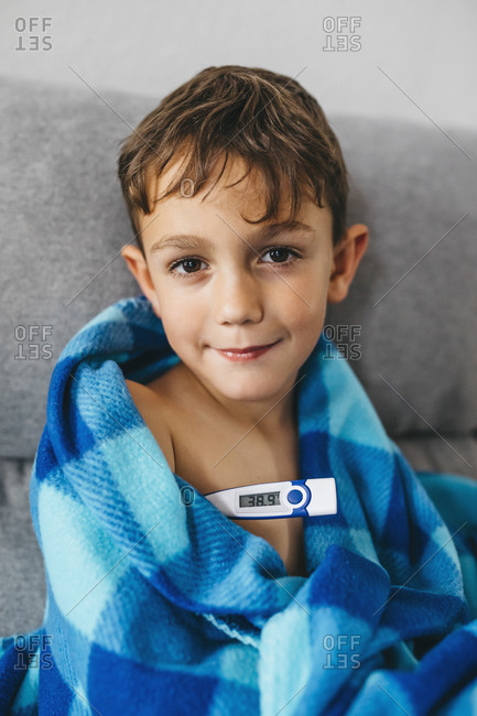 Portrait of sick boy sitting on couch with digital thermometer