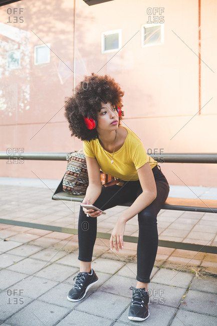 Young woman with afro hairdo listening to music with headphones at bus stop in the city