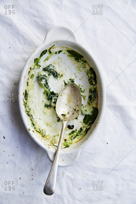 Empty white bowl with a spoon and bits of leftover spinach on a white countertop