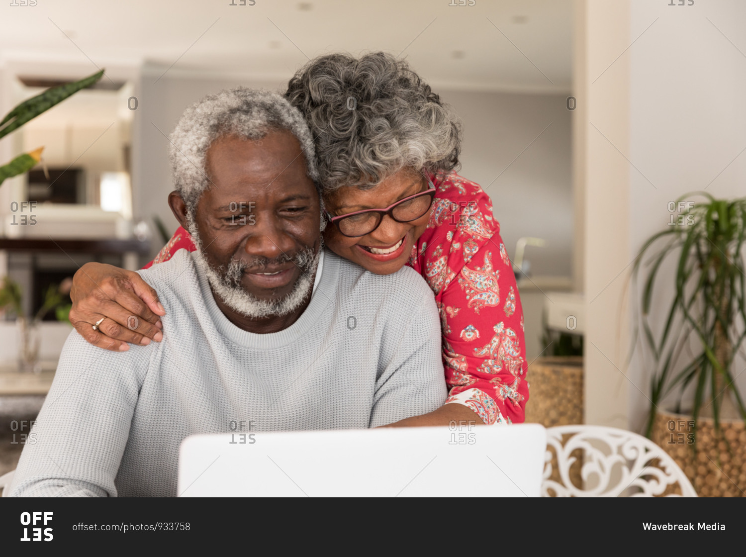 A senior African American couple spending time at home together, social distancing and self isolation in quarantine lockdown during coronavirus covid 19 epidemic, sitting at a table, using a laptop, embracing and smiling