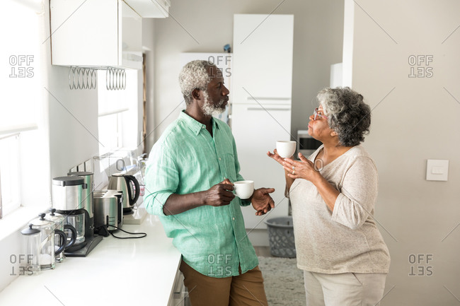 A senior African American couple spending time at home together, social distancing and self isolation in quarantine lockdown during coronavirus covid 19 epidemic, standing in the kitchen, talking, holding cups
