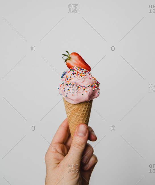 Hand holding strawberry ice cream cone against white background.