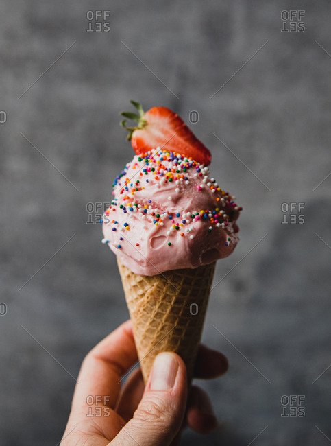 Hand holding strawberry ice cream cone against grey background.