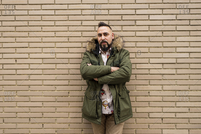 Bearded male with crossed arms leaning on bricked wall, looking camera