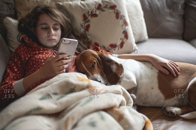 Tween girl looking at her phone while cuddling with hound dog on couch