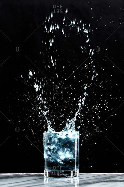 Ice creates a splash in a glass of blue liquid in a cocktail glass on a black background