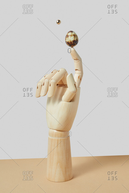 Wooden vertical standing dummy's hand with balancing quail egg on a finger and flying golden sphere near on a light duotone background, copy space