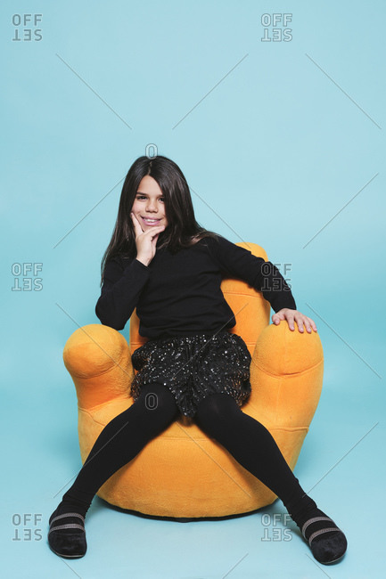 Preteen girl in black casual clothes resting hand on chin smiling looking at camera while relaxing in bright yellow armchair hand shape against light blue background in contemporary studio