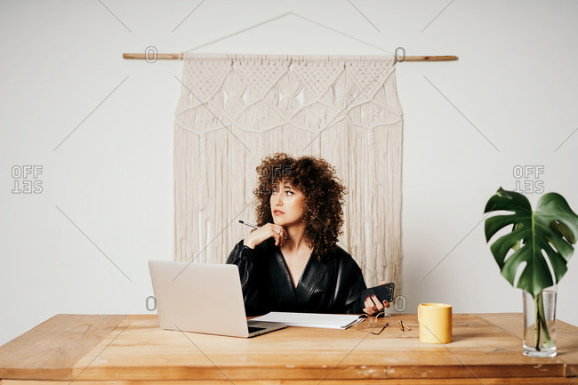 Adult lady in leather jacket and with curly hair sitting at table against macrame decoration and reading data on laptop during work on project in workplace