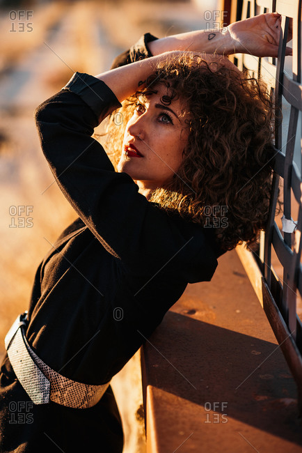 Side view of young woman with curly hair in trendy wear leaning on wall at city street in sunlight on blurred background