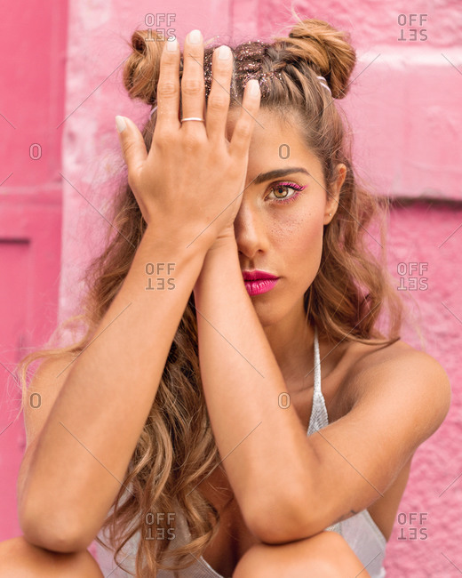 Glamour young woman with hair buns covering eye with hands and looking at camera while sitting against pink wall on city street