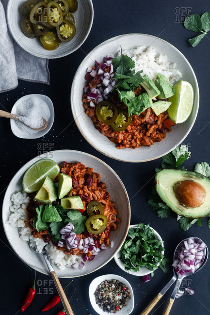 Top view of appetizing traditional Mexican chili with meat and white rice served in bowls with avocado and lime with chopped onion and marinated green chili pepper on black background with ingredients