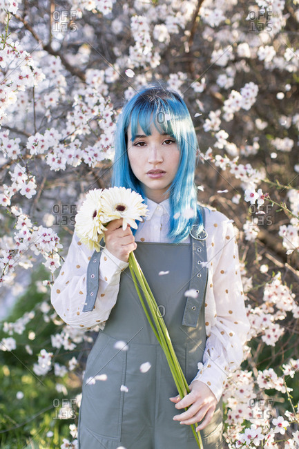 Modern trendy female with blue hair holding bouquet of fresh flowers and looking at camera while standing in blooming spring garden