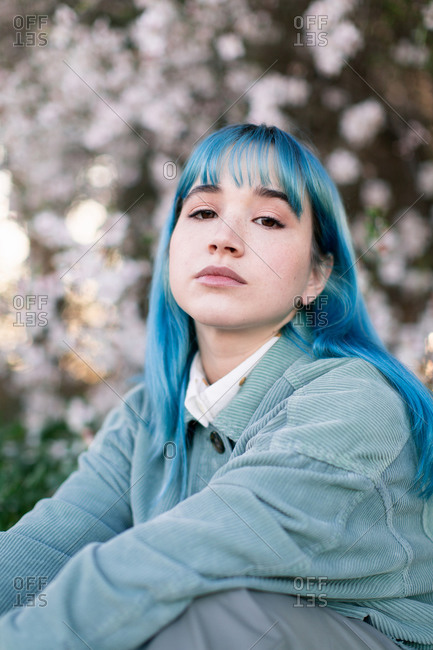 Sad millennial female model with blue hair in stylish outfit looking at camera thoughtfully while sitting on green grass near blooming tree in spring garden