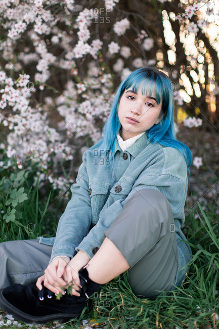 Sad millennial female model with blue hair in stylish outfit looking at camera thoughtfully while sitting on green grass near blooming tree in spring garden