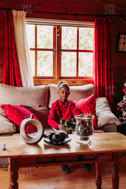 Satisfied black girl holding toy in hands and looking at camera while sitting on sofa near window in cozy living room with Christmas decoration