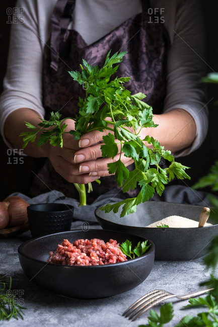 Unrecognizable female wearing apron keeping bunch of fresh parsley over bowl with raw minced meat while cooking lunch in kitchen