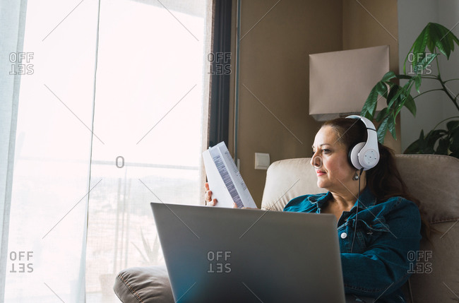 Mature woman listening to music in headphones and reading paper while sitting in armchair and doing remote job on laptop near window at home