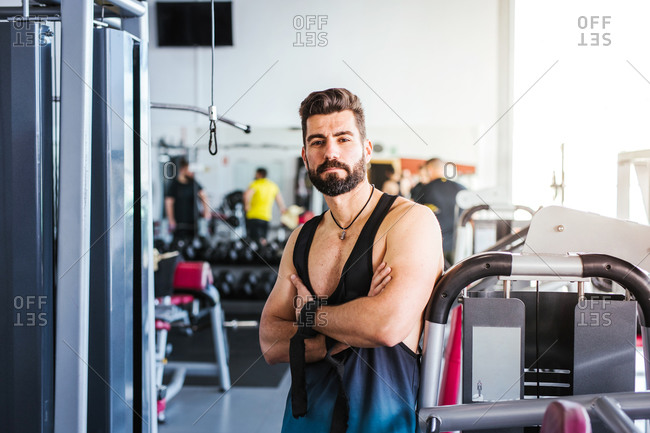 Muscular bearded man in sportswear looking at camera while standing with arms crossed and leaning on sports equipment in light contemporary gym