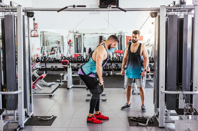 Confident instructor helping diligent client in sports clothes performing exercise on weight machine while standing against blurred interior of modern sport center