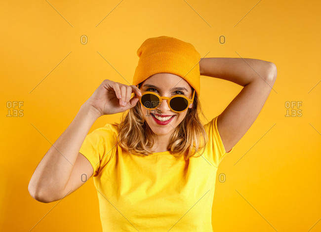 Happy young woman in bright clothes and stylish sunglasses smiling and looking at camera against orange background