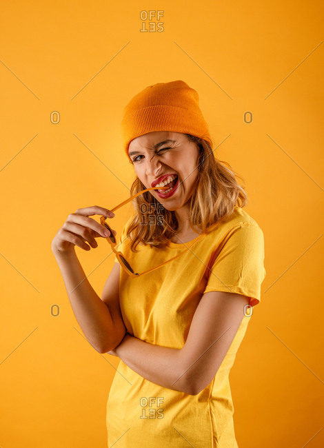 Side view of happy young woman in bright clothes and holding sunglasses to the mouth smiling and looking at camera while blinking an eyes against orange background