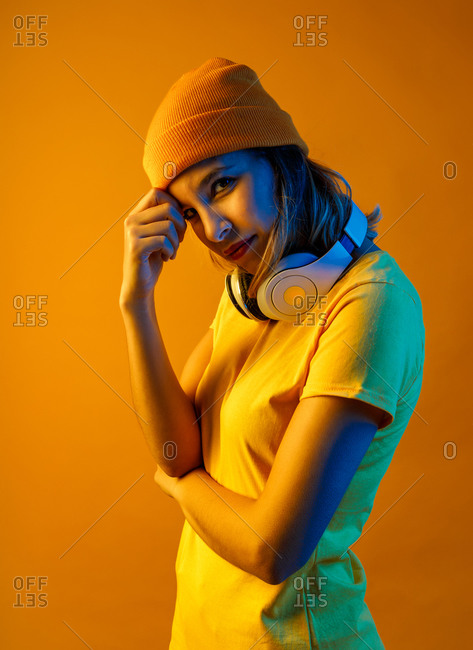 Thoughtful stylish young woman in orange beanie with headphones around the neck resting head over arm and looking at camera against orange background