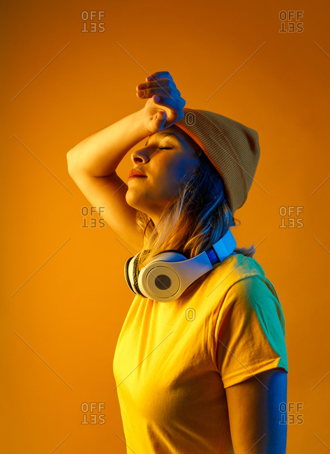 Thoughtful young woman in orange beanie with headphones around the neck keeping arm over head with closed eyes against orange background