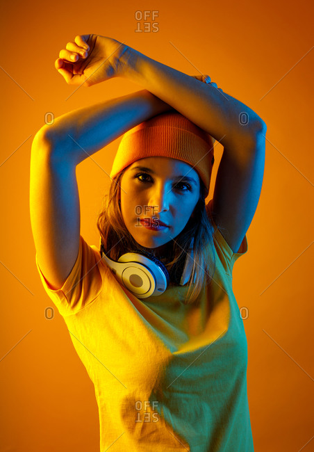 Confident stylish young woman in orange beanie with headphones around the neck keeping arm over head and looking at camera against orange background