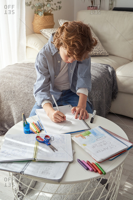 High angle of boy with curly hair sitting on sofa near round table and writing in notebook while doing homework in cozy living room at home