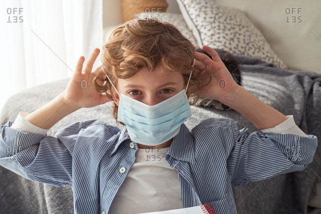 Dissatisfied boy in medical mask frowning and looking at camera while sitting near sofa with pet at home during quarantine